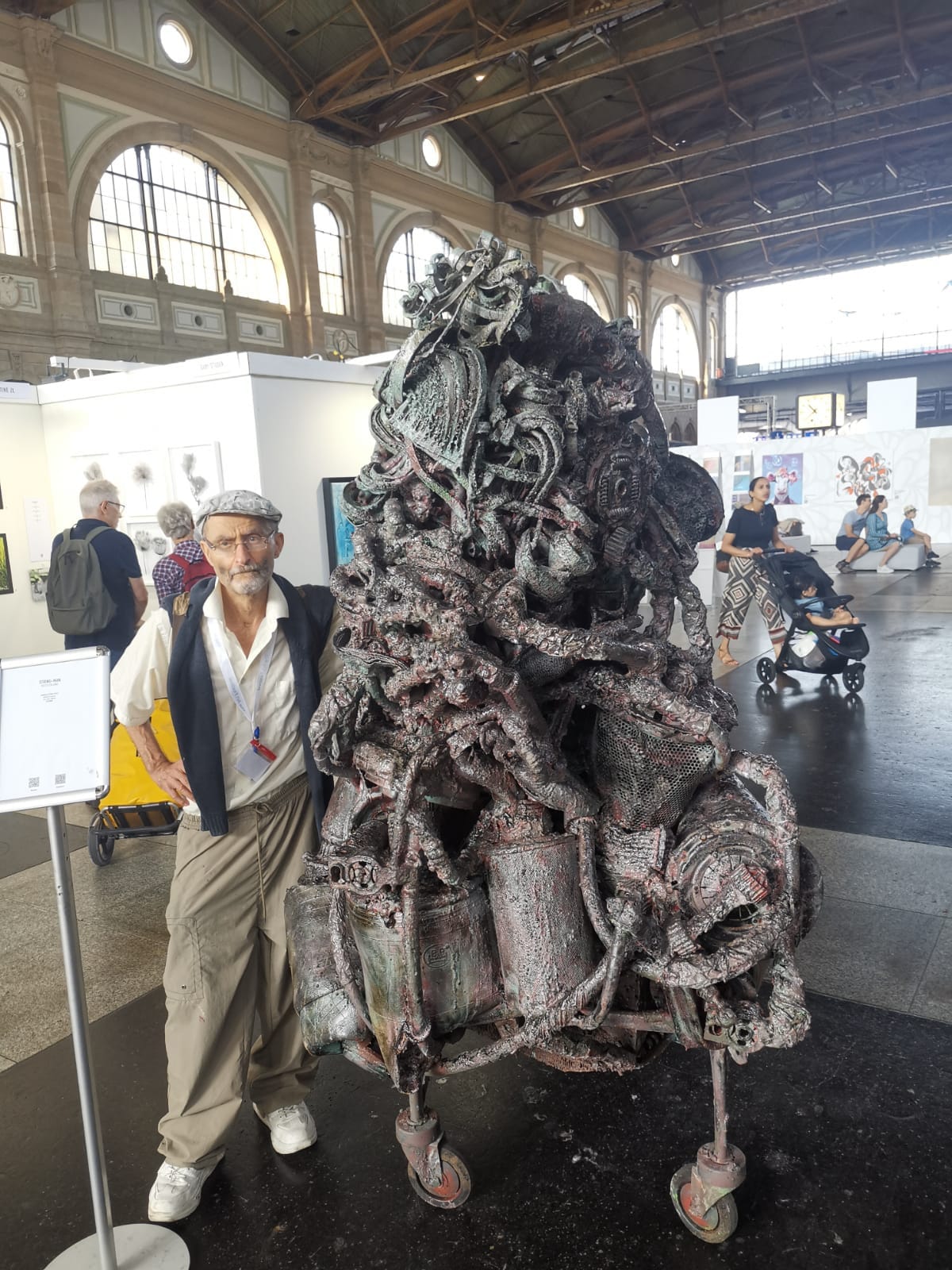 Hohe braun-graue Metallskulptur auf Rollen in einer großen Messehalle mit Bogenfenstern, ein Mann mit Schiebermütze steht daneben