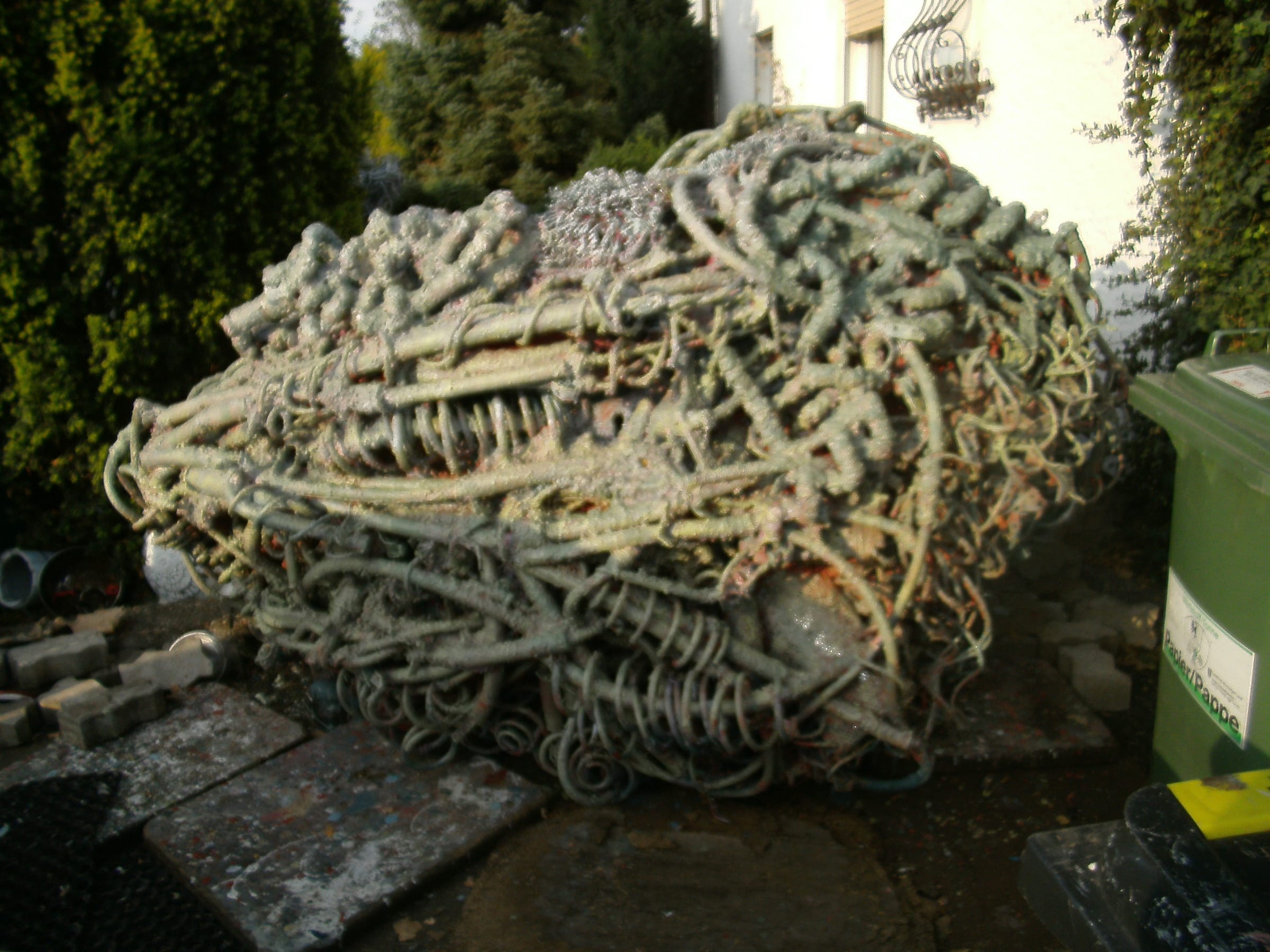 Liegende grün-graue Skulptur aus Metallschlingen im Hof, dahinter Hecke und Hauswand