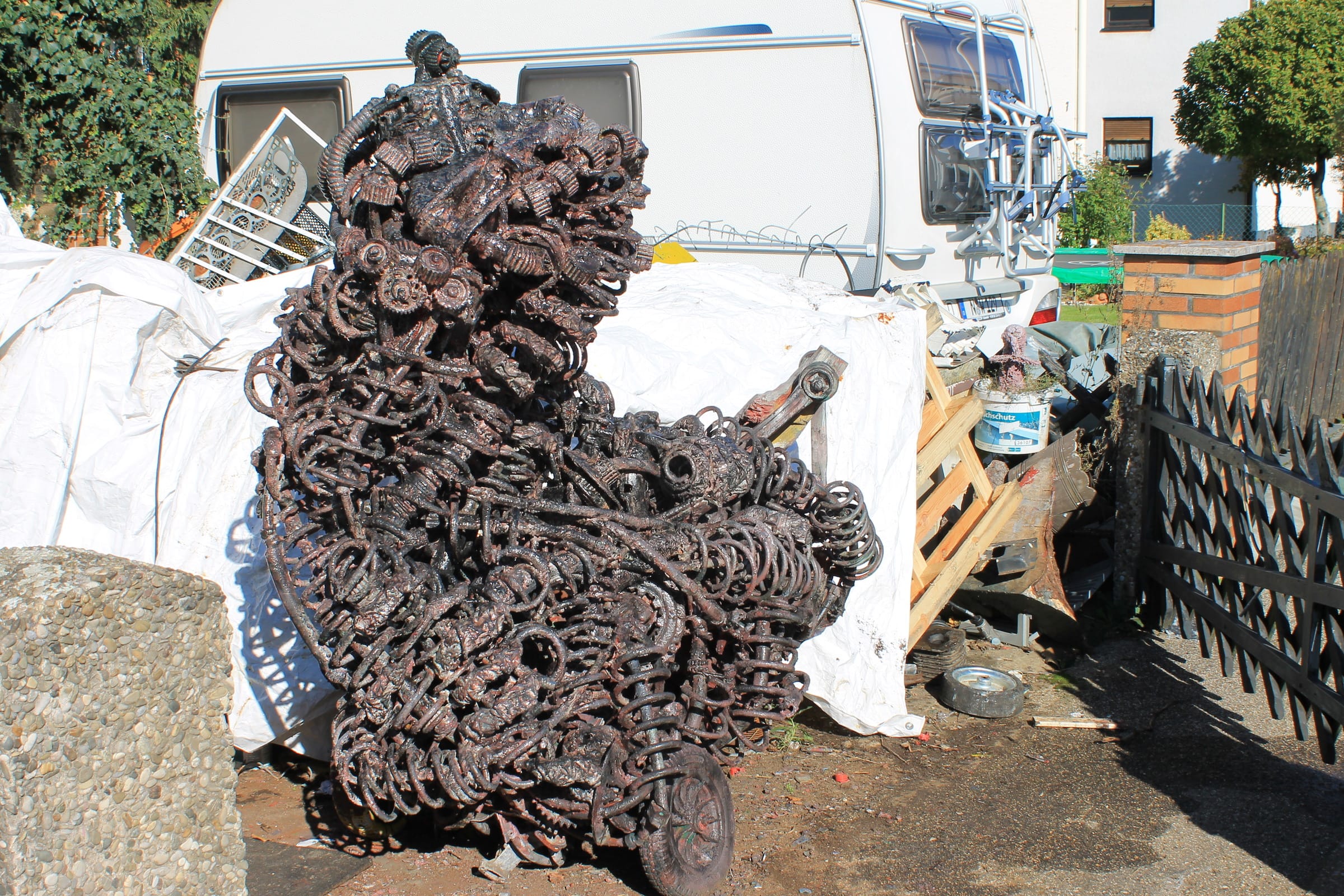 Kupferrote Metallskulptur aus Federn, Zahnrädern und Metallteilen auf Rollen, im Hof vor weißen Planen und einem Wohnwagen