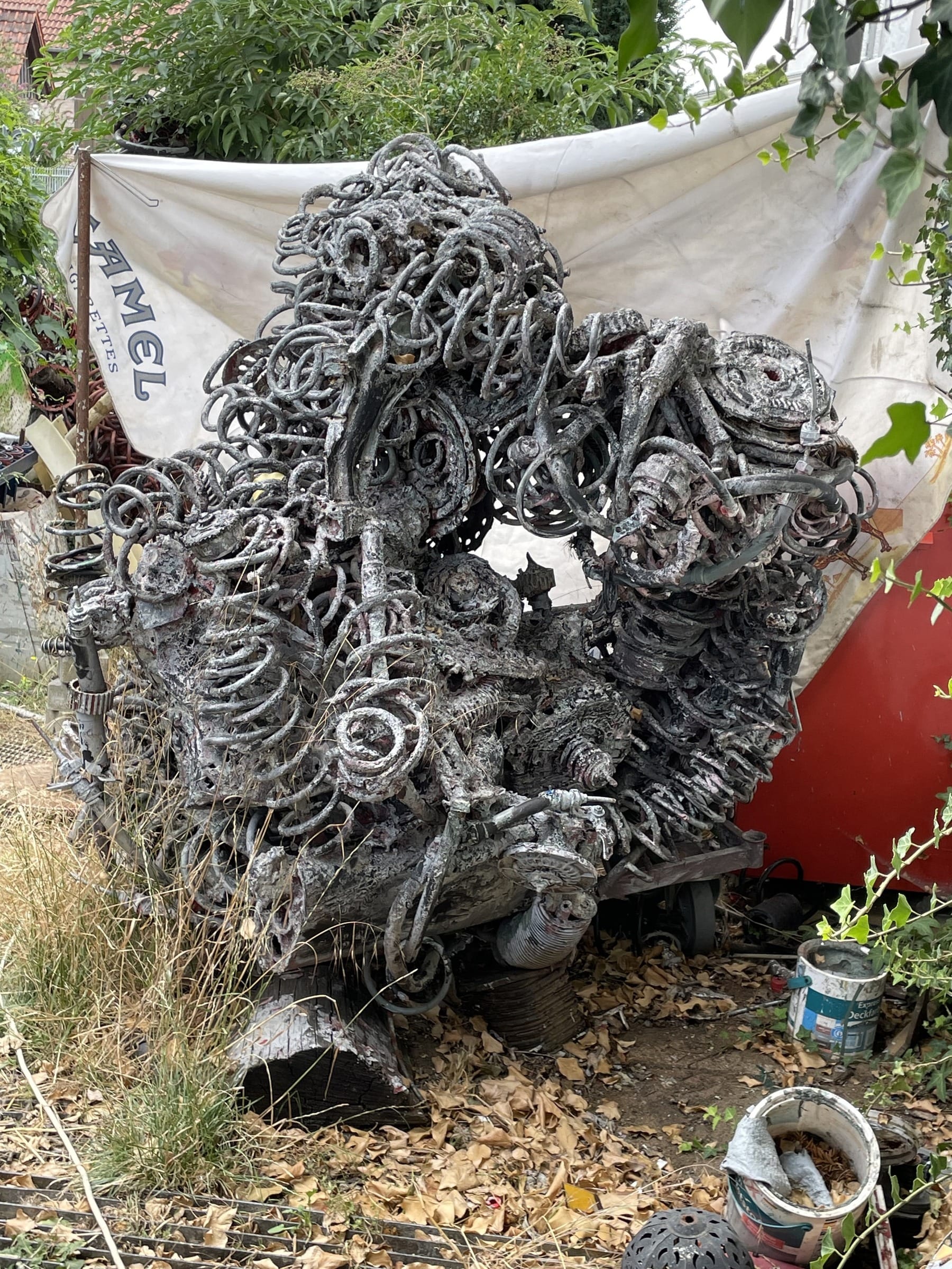 Silbergraue Skulptur aus Federn und Spiralen in einem verwilderten Garten vor einer weißen Plane