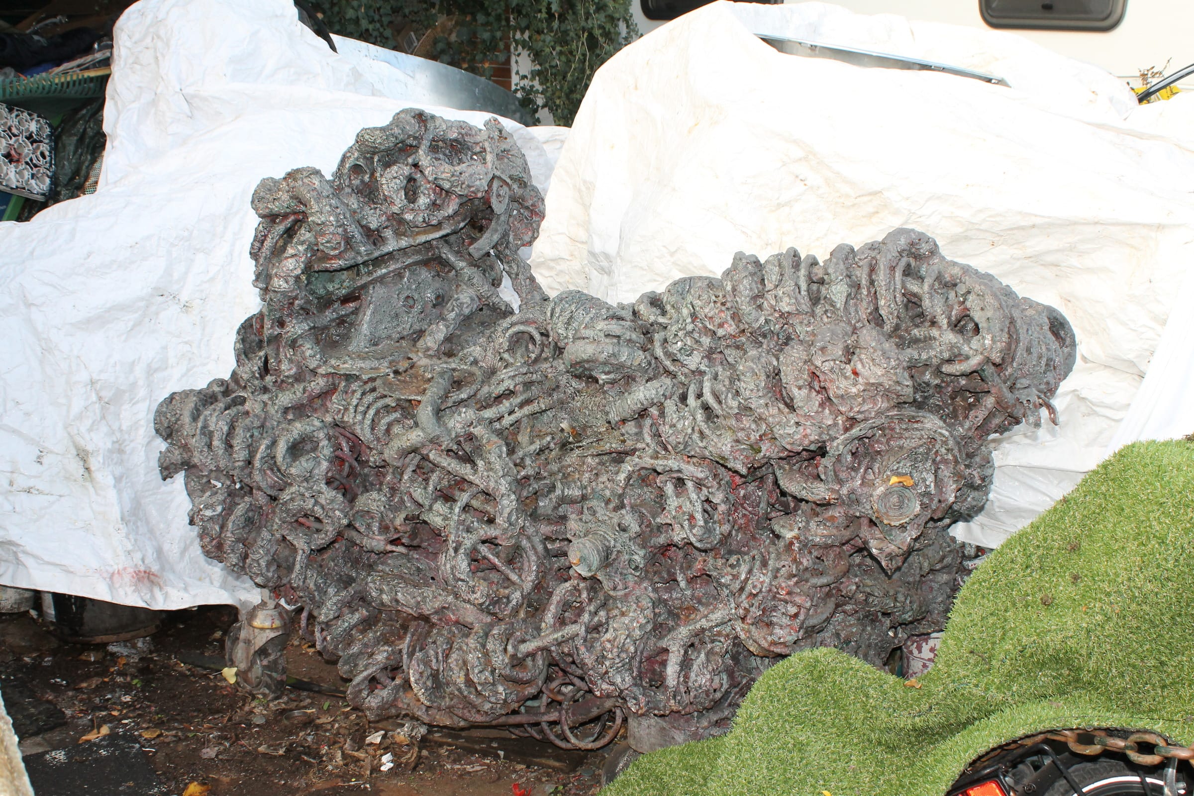 Breite grau-violette Skulptur aus Metallschlingen vor weißen Planen, rechts vorn Kunstrasen