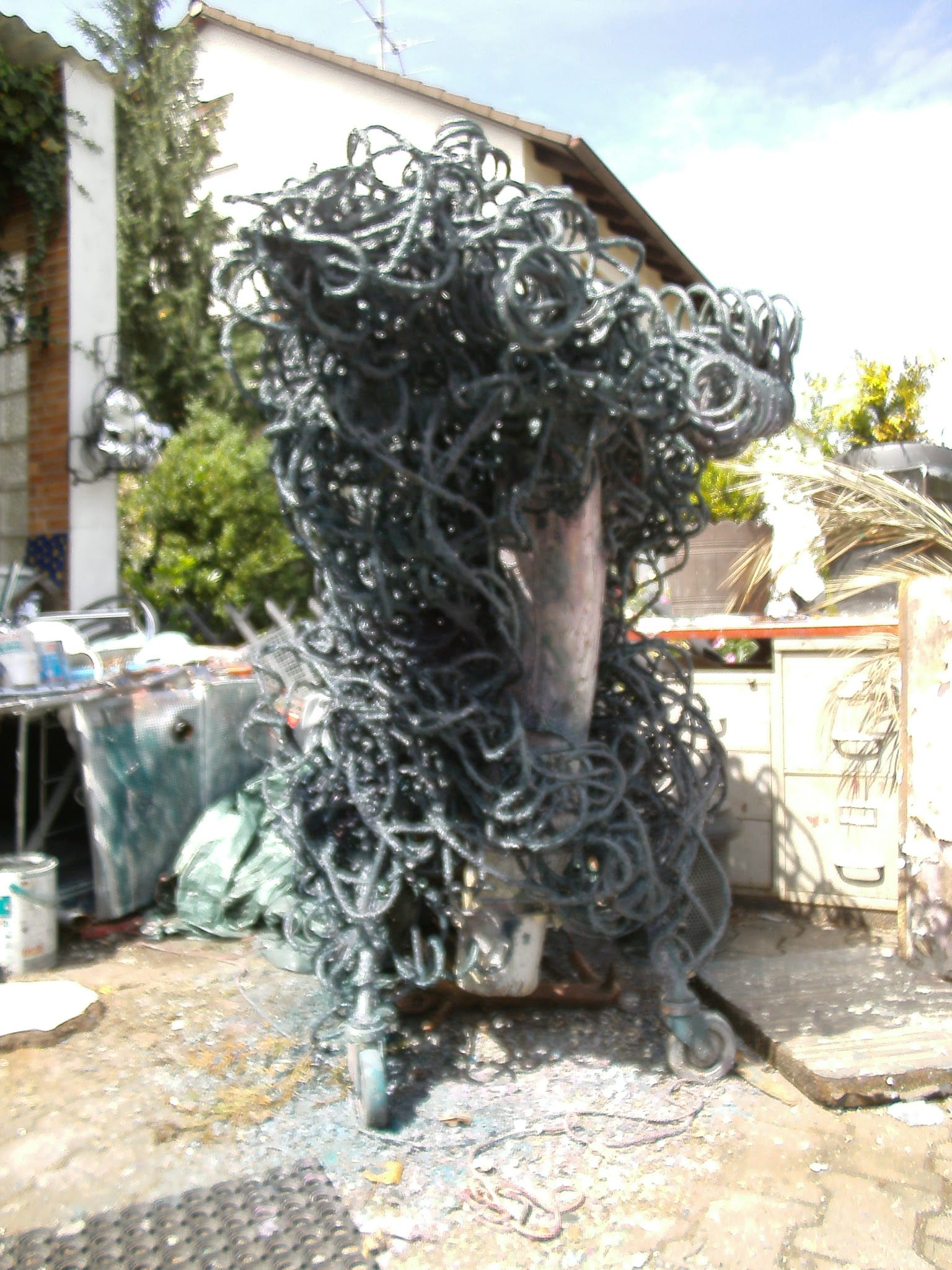 Hohe dunkel-grüngraue Skulptur aus Metallschlingen auf Rollen mit kupferfarbenem Kern, im Hof