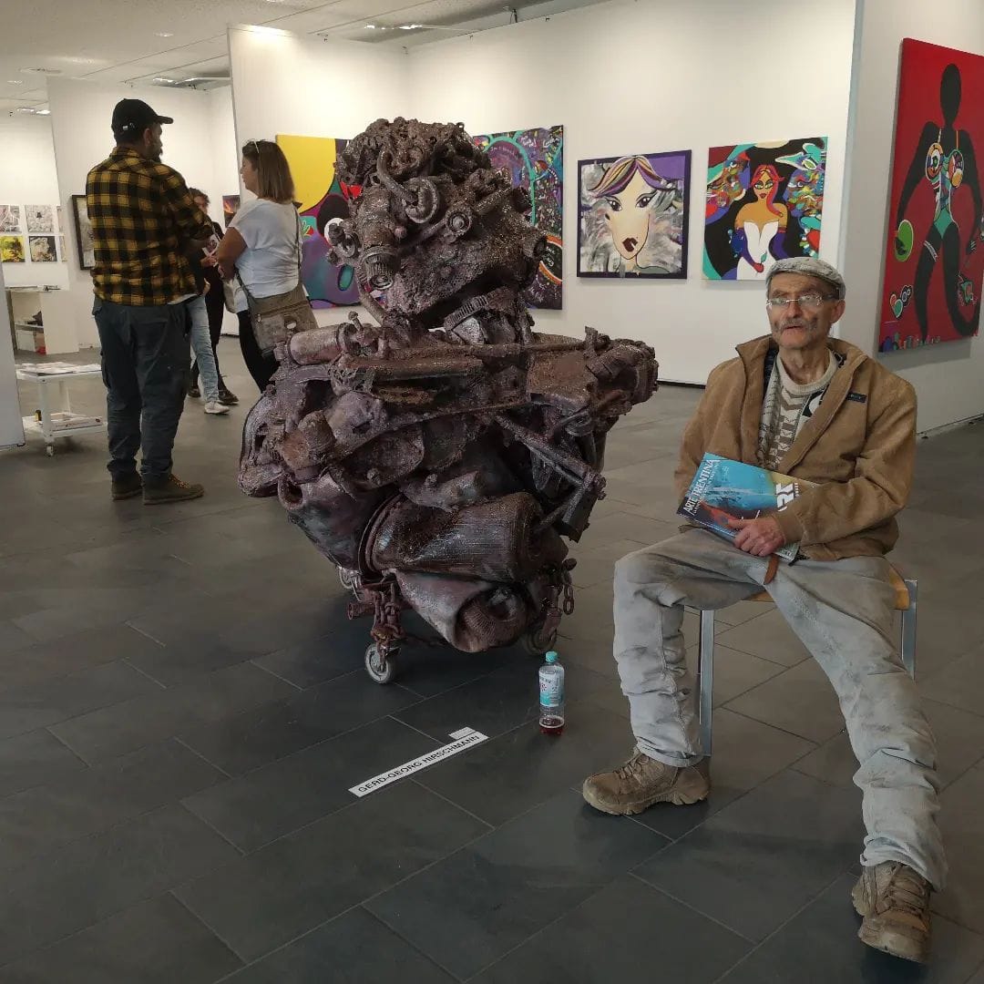 Braune Metallskulptur auf Rollen in einer weißen Galerie, der Künstler sitzt daneben, an der Wand Bilder anderer Aussteller